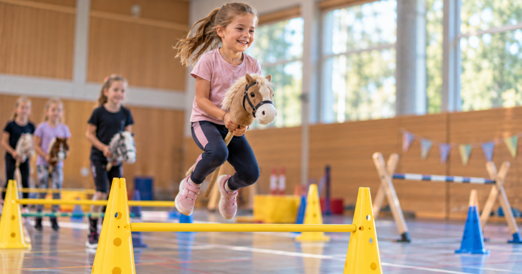 Mädchen springt lachend mit ihrem Hobby Horse über ein gelbes Hindernis in der Sporthalle